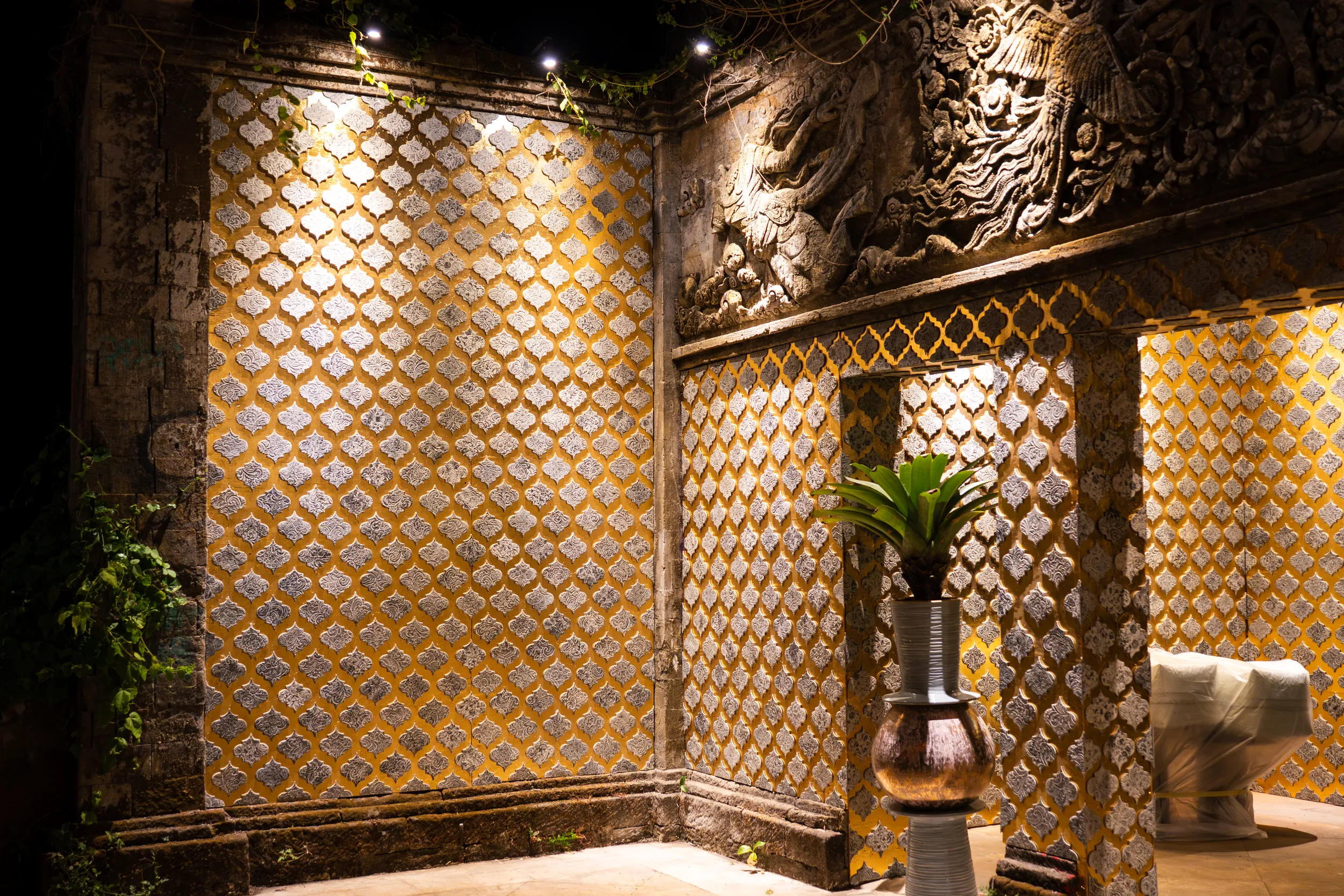 Wall of golden diamond-patterned ceramic tiles with a carved dragon relief and a plant in a copper vase